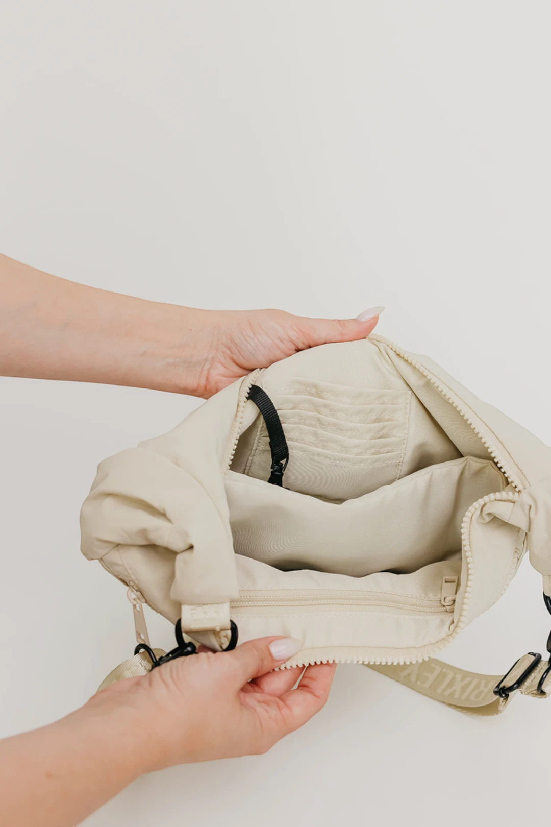 A close-up of a person's hands opening a beige nylon shoulder bag, showing the interior zippered compartments and black hardware against a plain white background.