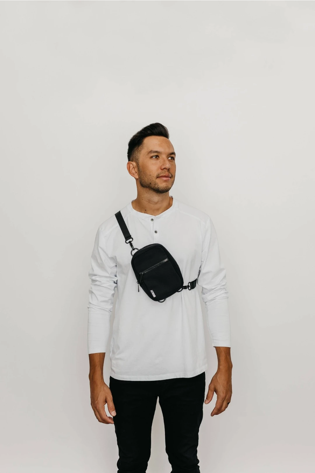 A man wearing a white long-sleeve henley shirt and black pants, wearing a black crossbody sling bag across his chest, standing against a plain white studio background.