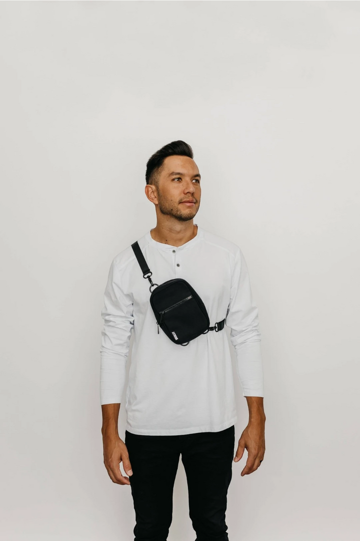 A man wearing a white long-sleeve henley shirt and black pants, wearing a black crossbody sling bag across his chest, standing against a plain white studio background.