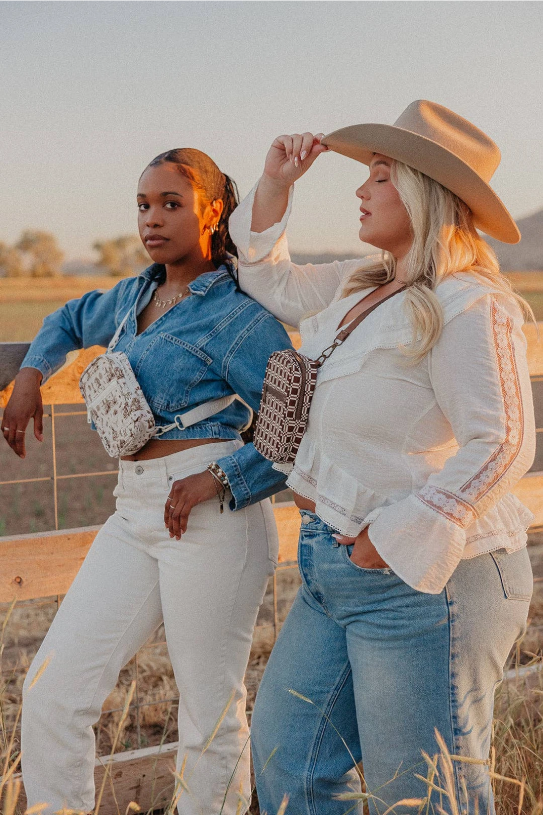Two women posing outdoors in a field at sunset. One wears a denim jacket, white pants, and a white crossbody bag, while the other wears a white embroidered blouse, blue jeans, and a beige cowboy hat, both carrying crossbody bags.