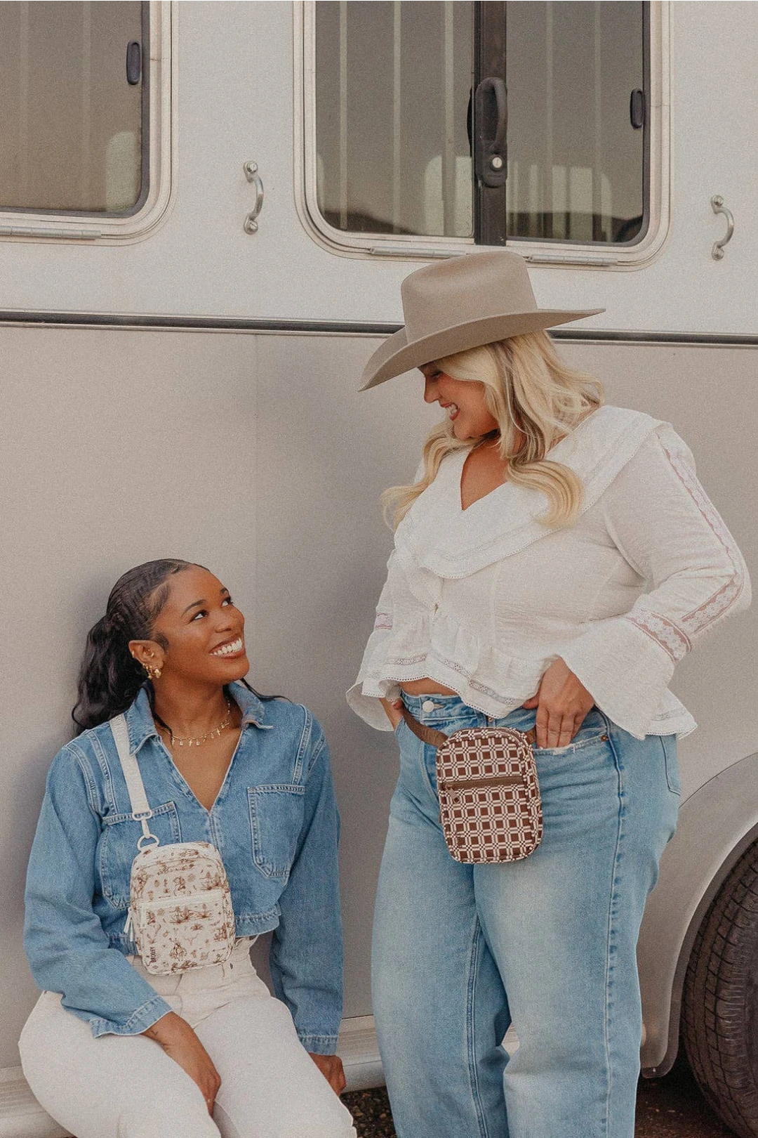 Two women posing in front of a vintage-style white RV. The woman on the left is wearing a light blue denim jacket, white pants, and a white floral crossbody bag.