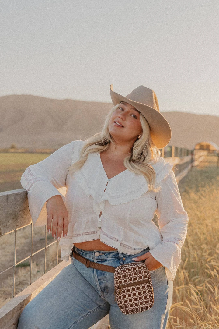 A woman posing outdoors in a rural setting, wearing a white ruffled long-sleeve crop top, light blue jeans, a brown belt, and a beige cowboy hat. She is leaning against a wooden fence, with a small brown and beige patterned crossbody bag hanging from her waist.