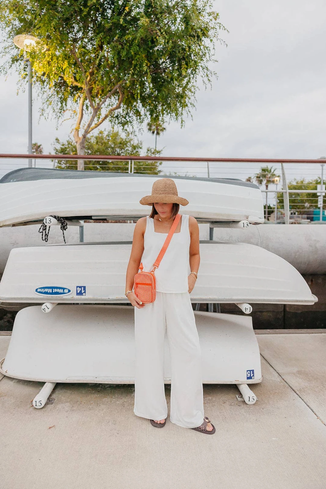 A woman stands outdoors in front of stacked white boats, wearing a straw hat, a sleeveless white outfit, and carrying a small orange crossbody bag.