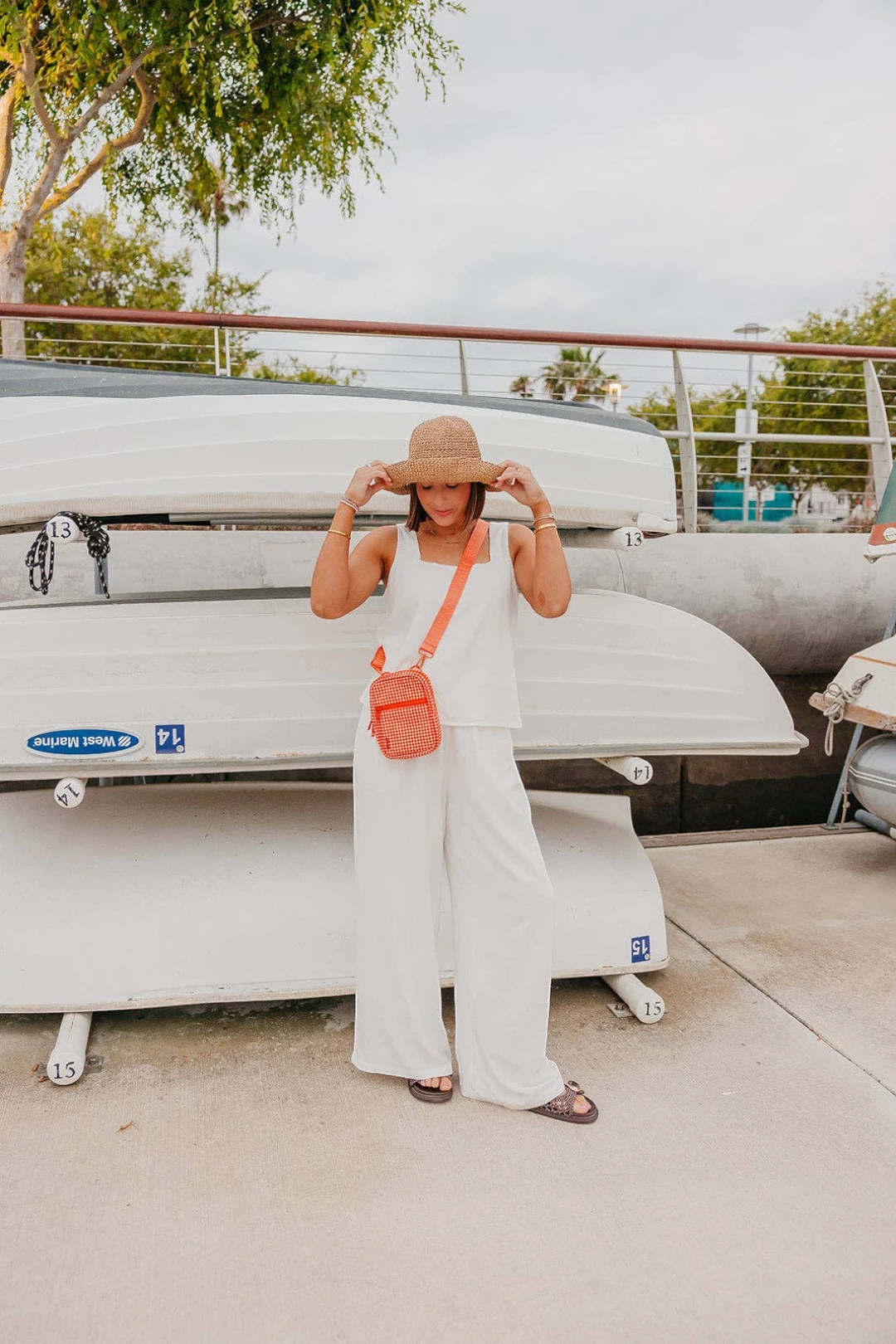 A woman stands outdoors in front of stacked white boats, wearing a sleeveless white outfit with wide legs, a tan hat, and a small orange crossbody bag.
