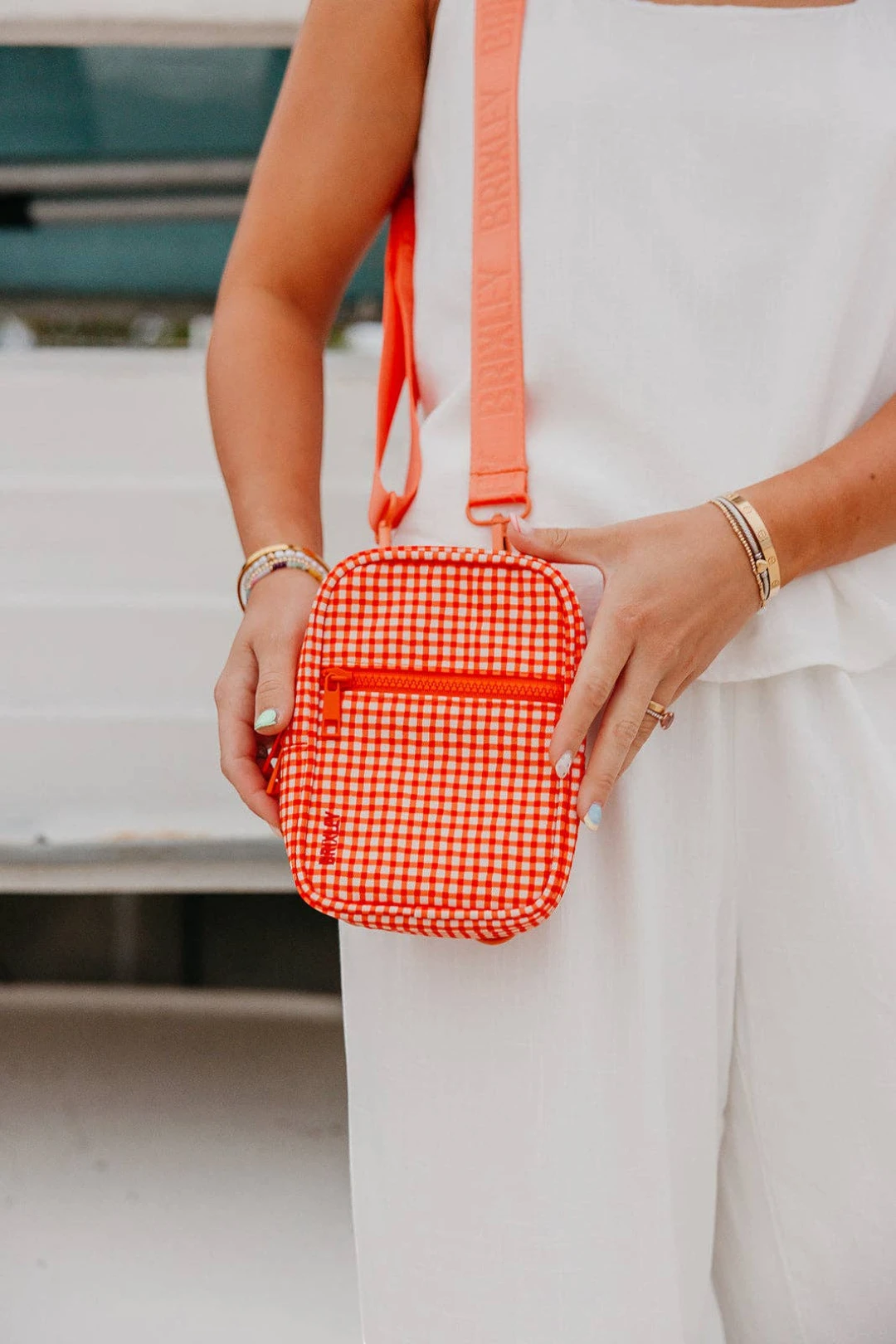 A person in a white sleeveless outfit holds a small red-and-white gingham crossbody bag with a bright coral-orange strap and trim. The photo is cropped from the chest down, with the person's hands and arms visible against a softly blurred outdoor background.