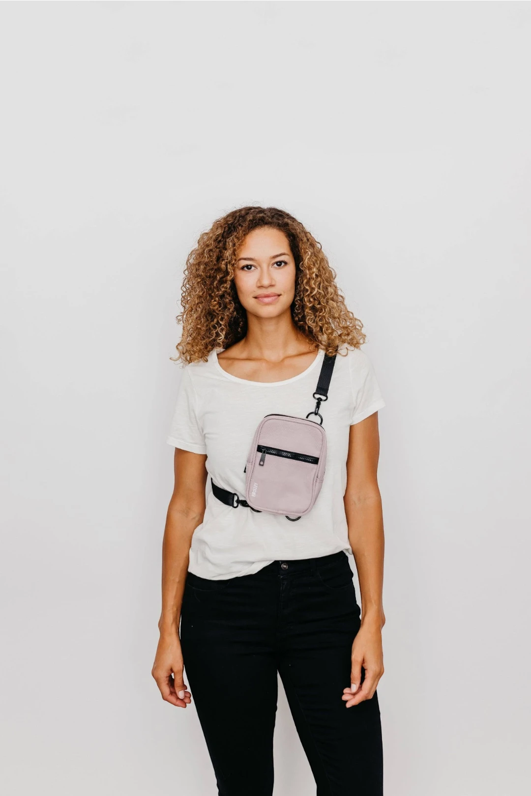 A woman with curly hair wearing a white t-shirt and black pants models a small, dusty pink crossbody bag with black straps and trim against a plain white background.