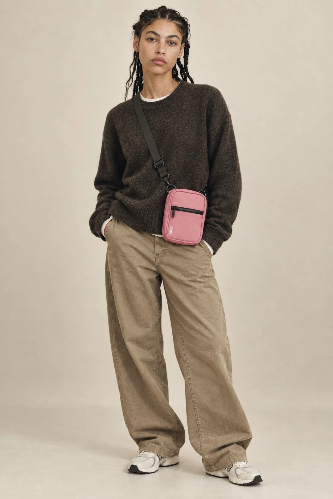 A model stands against a plain beige studio background wearing a dark brown knit sweater, beige wide-leg pants, white sneakers, and a small pink crossbody bag with a black strap.