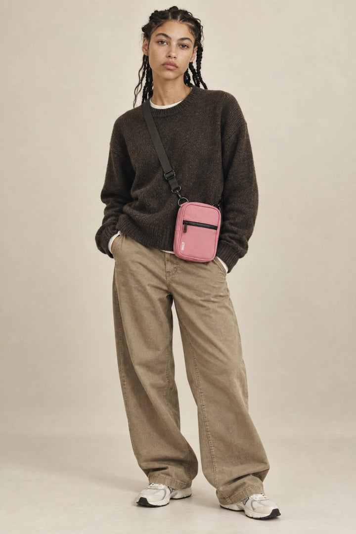 A model stands against a plain beige studio background wearing a dark brown knit sweater, beige wide-leg pants, white sneakers, and a small pink crossbody bag with a black strap.