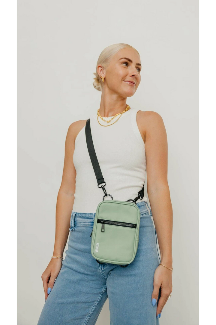 A woman wearing a white ribbed tank top and light blue jeans models a sage green crossbody phone bag with black trim and strap against a plain light gray background.
