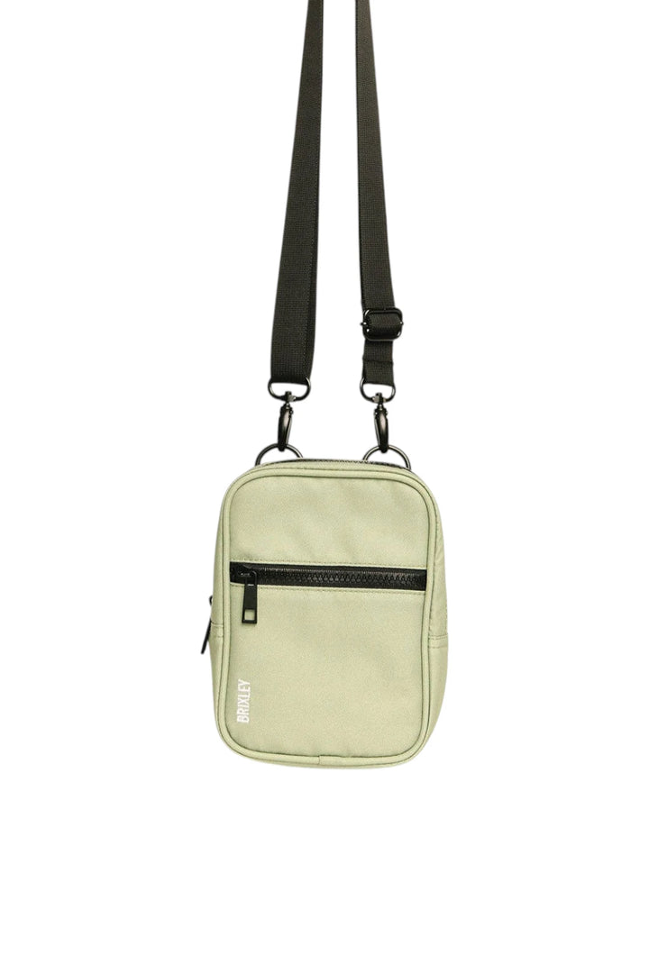 A light sage green crossbody bag with a black adjustable shoulder strap, black zipper, and a small white vertical logo on the front pocket, shown against a plain white background.