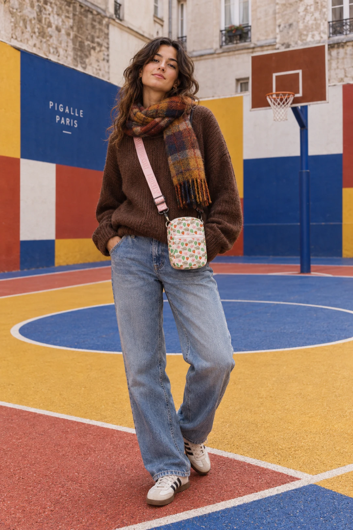 A woman stands on a colorful outdoor basketball court wearing a brown fuzzy sweater, a plaid scarf, light blue jeans, white sneakers, and carrying a small crossbody bag with a pink strap.
