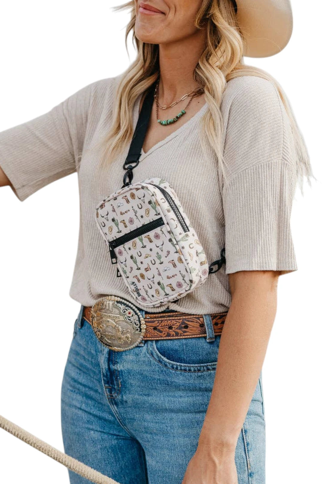 A woman wearing a small crossbody bag with a white background and a colorful western-themed pattern of horseshoes, boots, and buckles. She is also wearing a beige ribbed top, blue jeans, a brown leather belt with a large silver buckle, and a cowboy hat, set against a plain white background.
