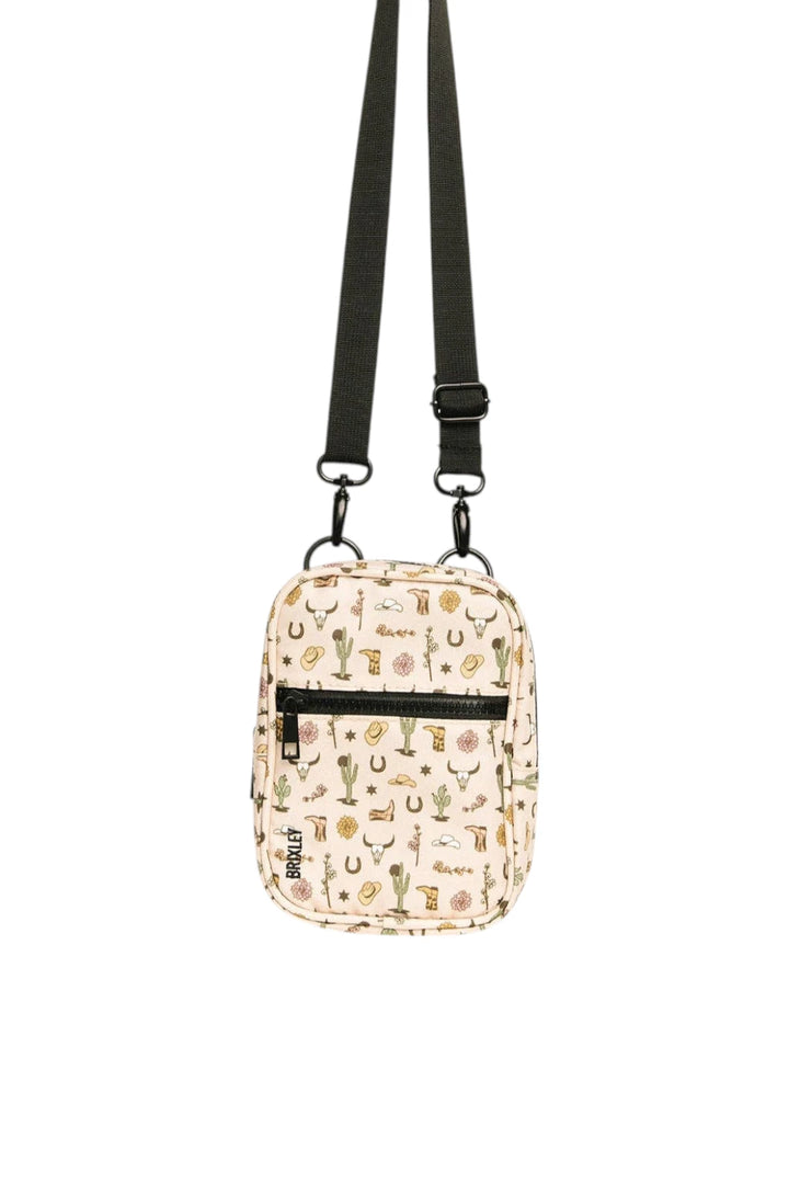 A small crossbody bag with a beige background and a colorful pattern of cacti, horseshoes, and western-themed illustrations. It features a black adjustable shoulder strap, a black front zipper pocket, and a black vertical brand tag that reads 'trtl'.