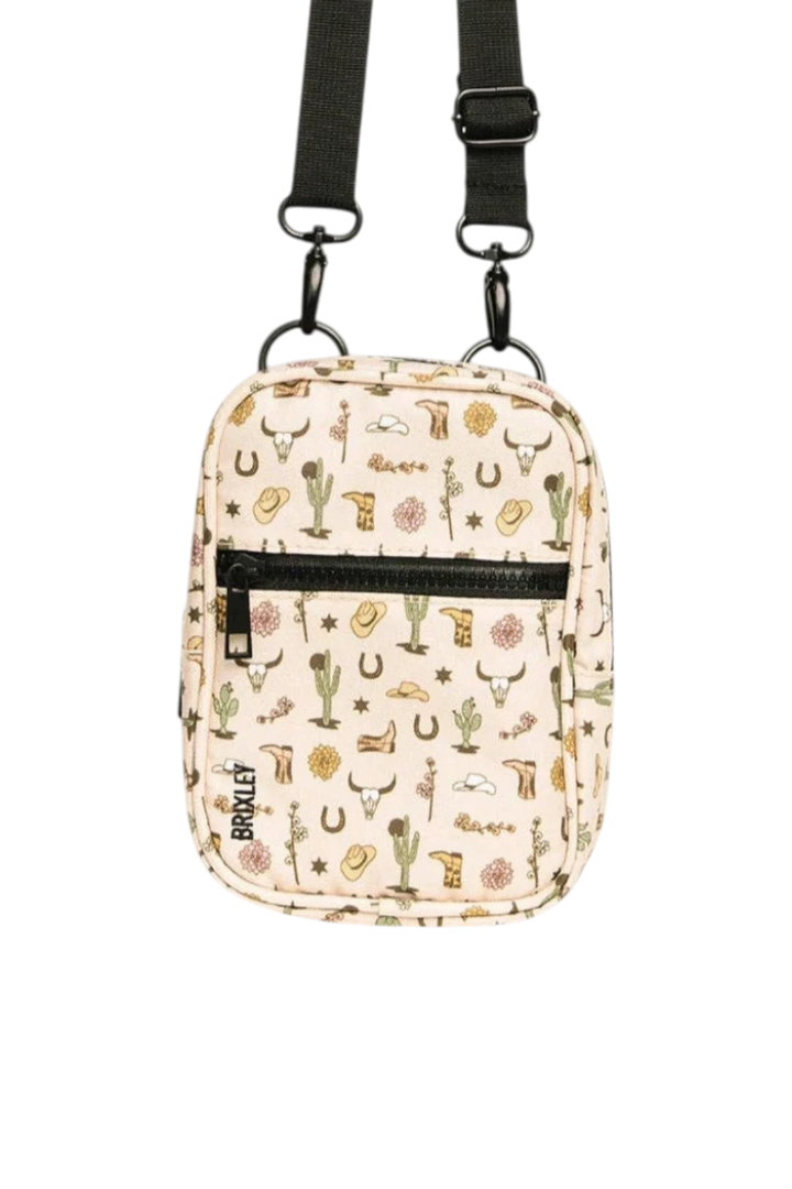 A small crossbody bag with a western-themed print on a cream background, featuring a black zipper, black strap, and western motifs like boots, horseshoes, and cacti. The brand name 'BRIXLEY' is printed vertically on the front.