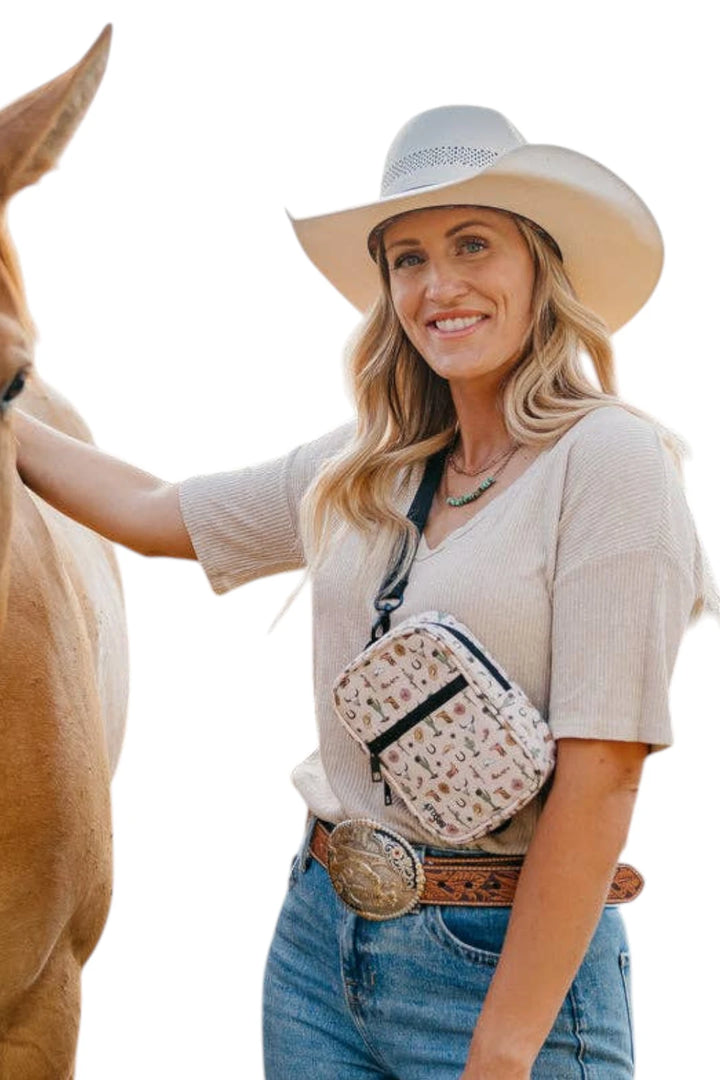 A woman wearing a cream cowboy hat, beige short-sleeve top, and blue jeans stands beside a horse, wearing a small beige crossbody bag with a western pattern and black trim.