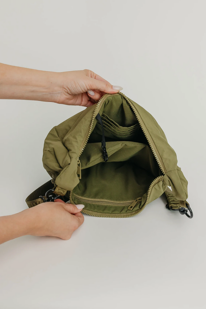 An olive green nylon crossbody bag is shown open, with a person's hands holding the top edge to display the interior compartments, zipper, and drawstring closure against a plain light background.