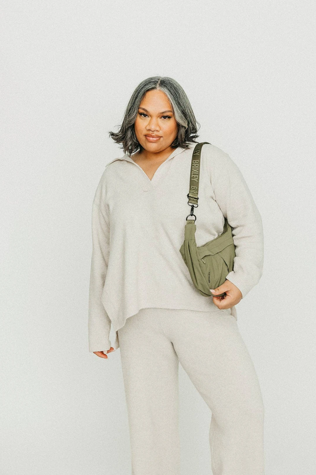 A woman modeling a light beige knit lounge set consisting of a half-zip pullover and matching sweatpants, carrying an olive green crossbody bag with a visible 'BRIXLEY' brand strap, set against a plain light gray studio background.