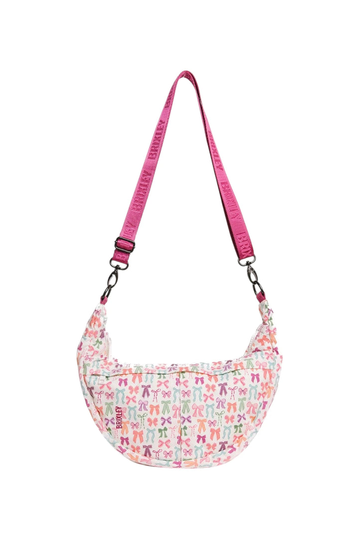 A crescent-shaped shoulder bag with a white background covered in a colorful pattern of bow ties, featuring a bright pink adjustable strap with 'BRIXLEY' printed repeatedly.