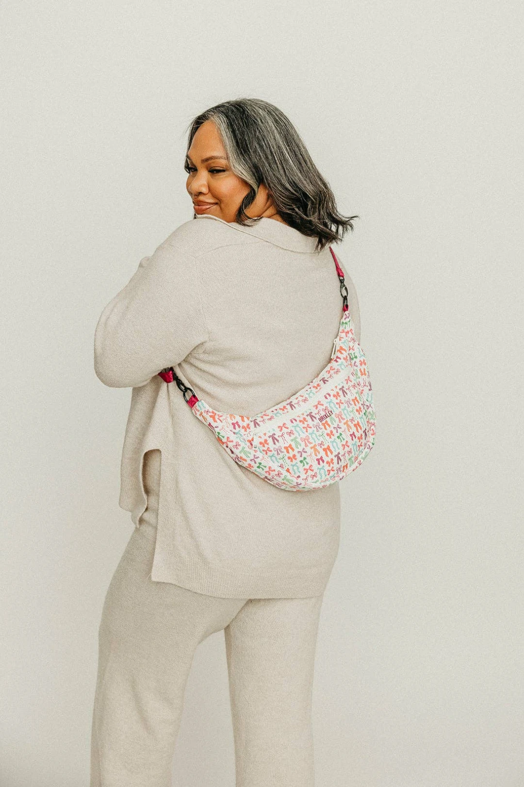 Person wearing a beige suit with a bow patterned sling bag on a plain background