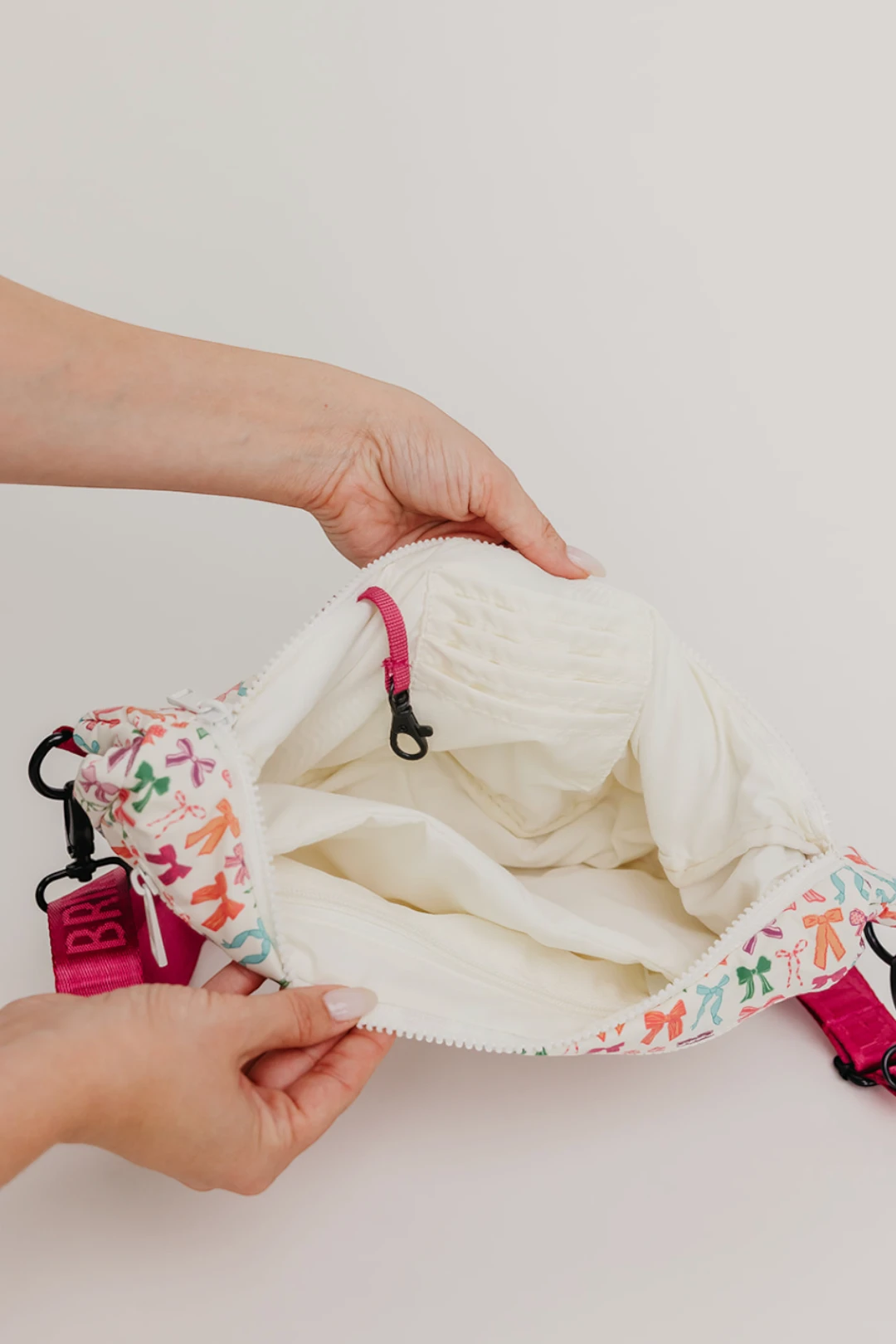 A close-up of a person's hands opening a cream-colored soft bag with a colorful bow-patterned fabric trim and bright pink straps, set against a plain off-white background.