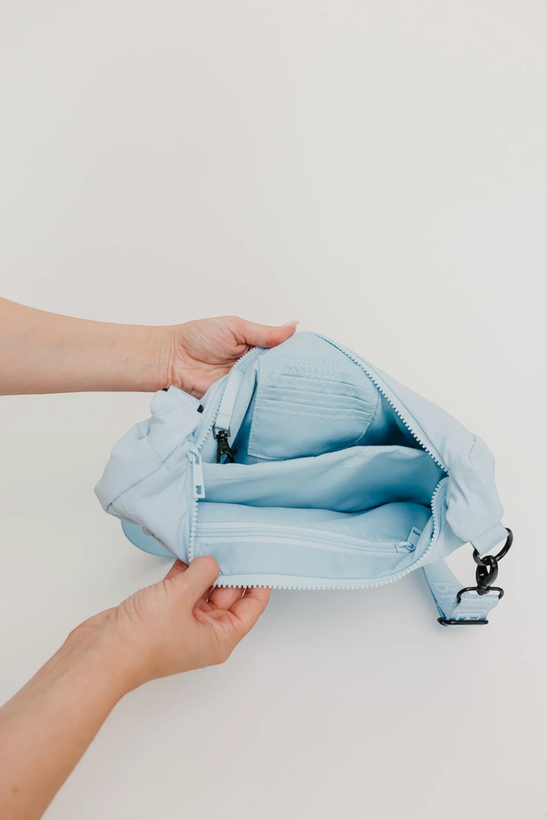 A light blue crossbody bag with a zippered main compartment and multiple interior pockets, held open by two hands against a plain off-white background.