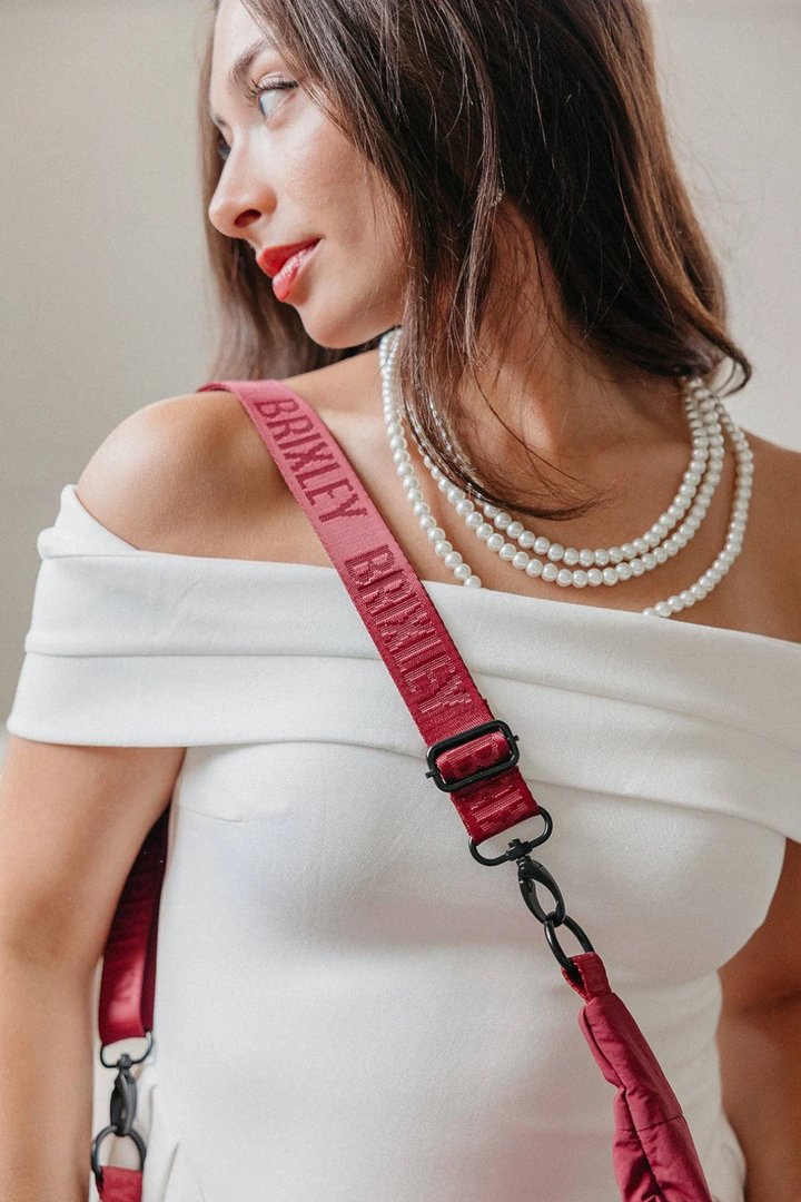 A close-up lifestyle shot of a woman wearing a white off-the-shoulder top and layered pearl necklaces, wearing a prominent burgundy leather crossbody bag strap with 'BRIXLEY' embossed on it.