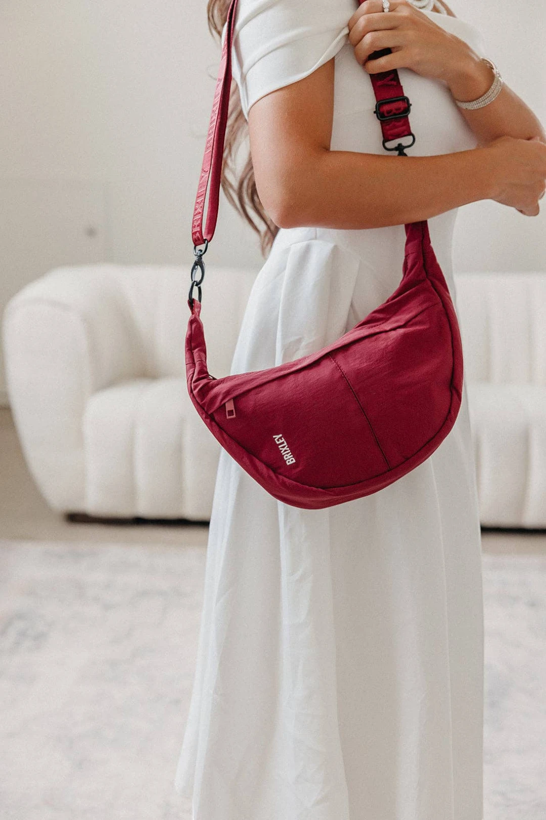 A woman in a white dress wearing a red crescent-shaped shoulder bag with a matching strap, standing in a bright room with a white sofa in the background.