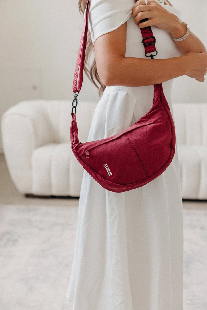 A woman in a white dress wearing a red crescent-shaped shoulder bag with a matching strap, standing in a bright room with a white sofa in the background.