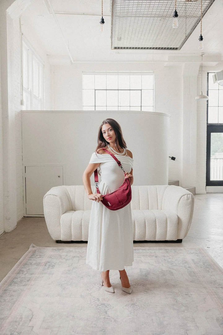 A woman wearing a white off-the-shoulder dress and white pointed-toe heels stands in a bright, modern white loft studio, carrying a deep burgundy crossbody bag with a red strap.