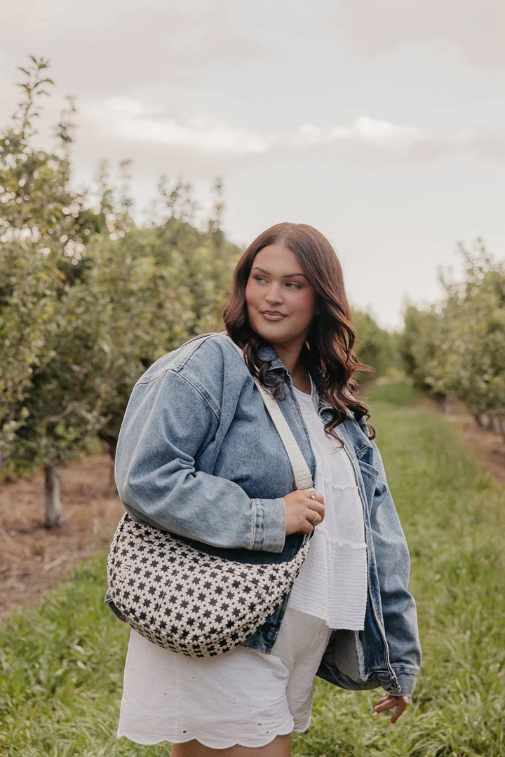 A woman standing in an apple orchard wearing a denim jacket, white ruffled top and skirt, and carrying a black and white patterned shoulder bag.
