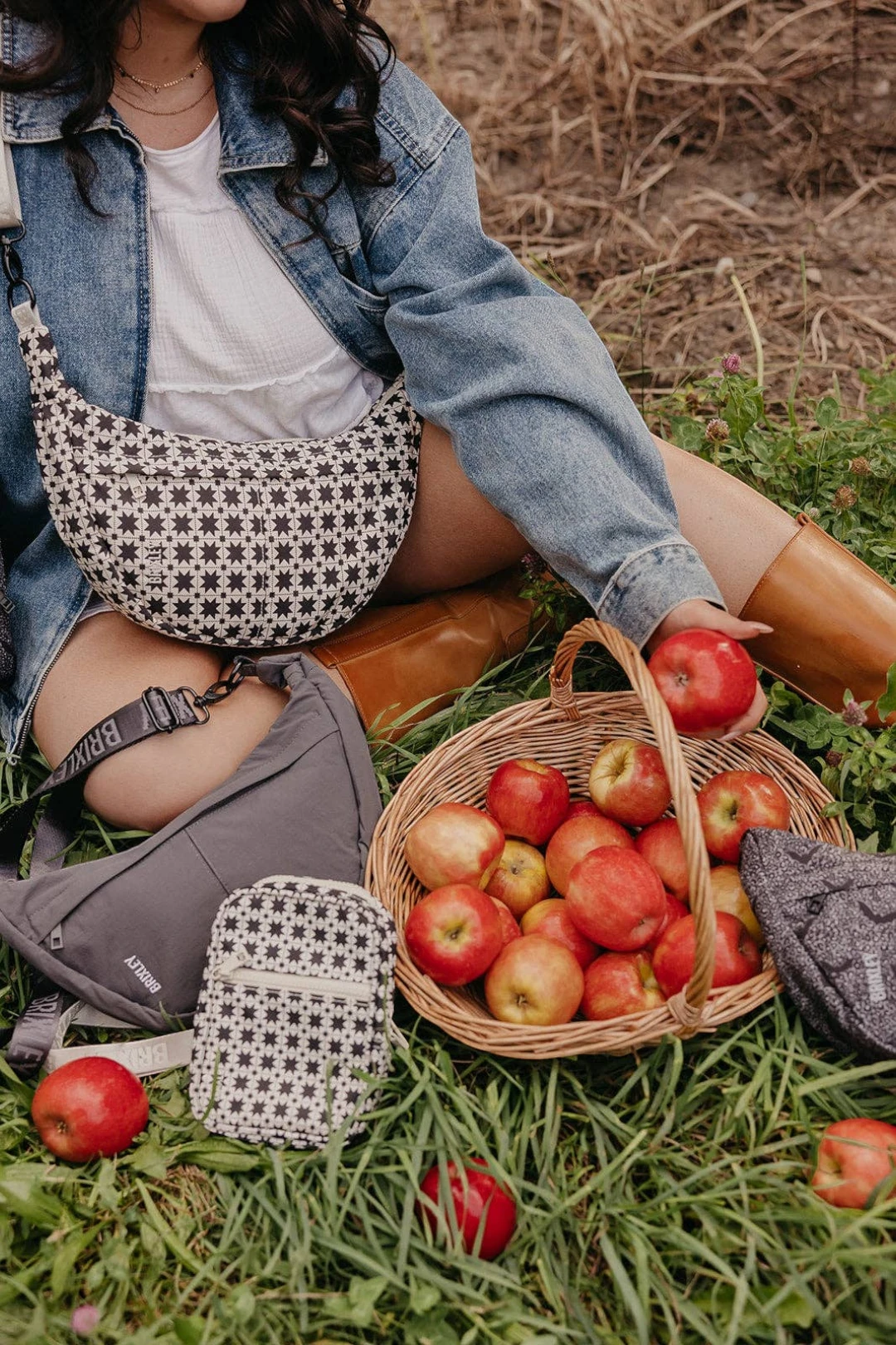 A person sitting outdoors in grass next to a basket of red apples, wearing and carrying multiple patterned crossbody bags in a rustic outdoor setting.