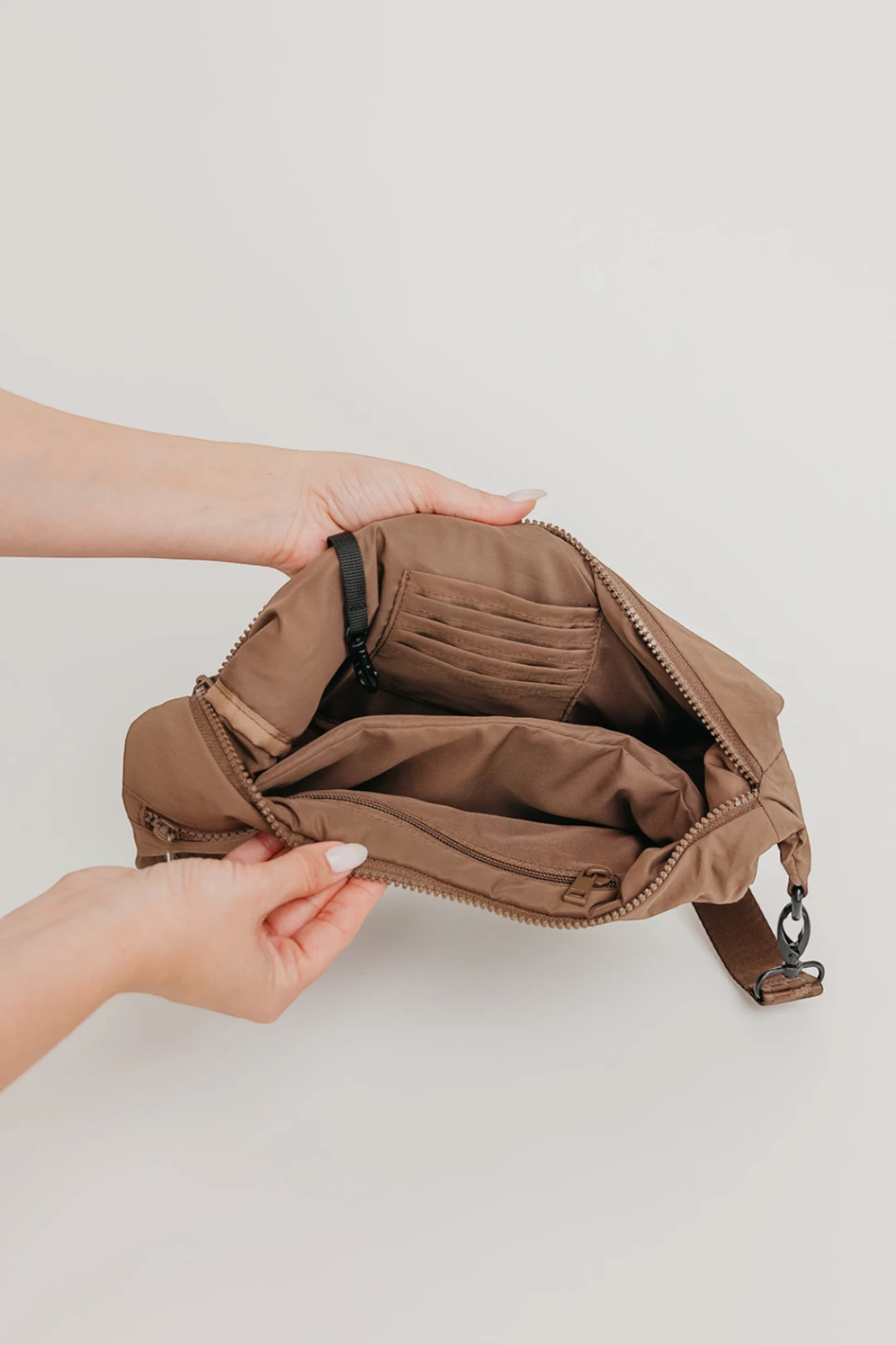 A brown crossbody bag with multiple interior compartments, shown open and held by two hands against a light gray background.
