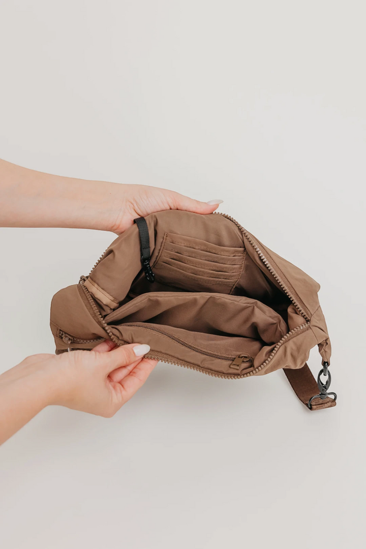 A brown crossbody bag with multiple interior compartments, shown open and held by two hands against a light gray background.