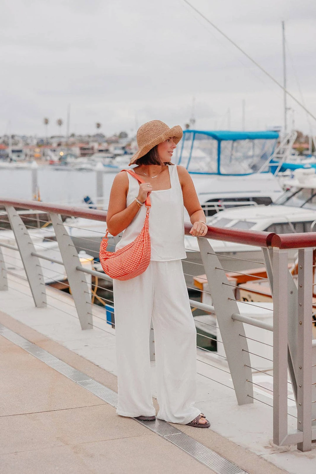 A woman stands on a marina walkway wearing a white sleeveless top and matching wide-leg pants, a beige straw hat, and carrying a coral-orange woven shoulder bag.
