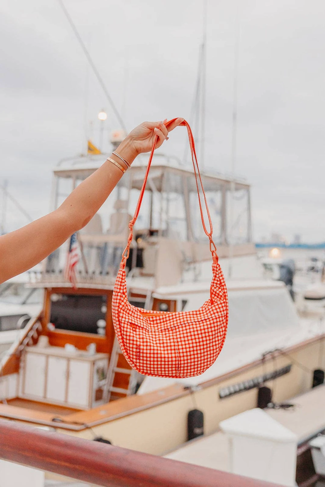 A person holds up a red and white gingham crescent-shaped shoulder bag with a long strap, set against a blurred background of boats on the water.