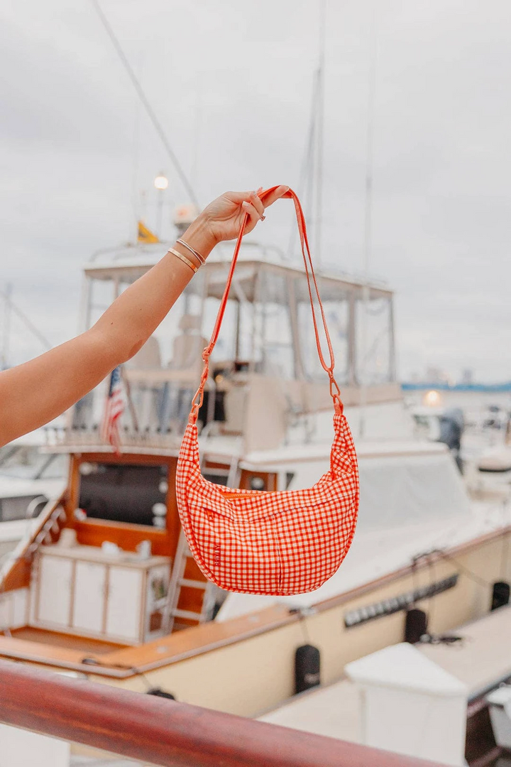 A person holds up a red and white gingham crescent-shaped shoulder bag with a long strap, set against a blurred background of boats on the water.