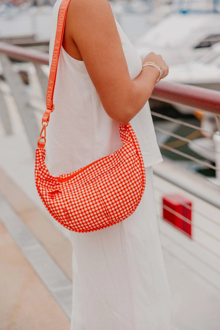 A woman in a white dress carries a crescent-shaped shoulder bag with a red and white gingham pattern and a matching red strap, set against a blurred background of boats and water.