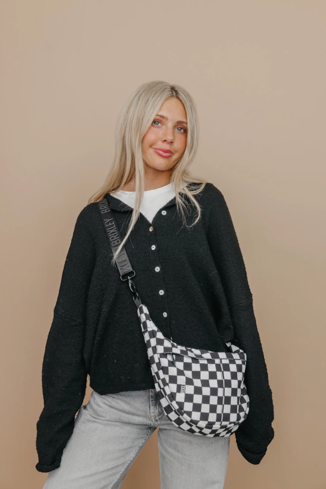 Woman wearing a black cardigan and light gray pants with a checkered bag on a beige background