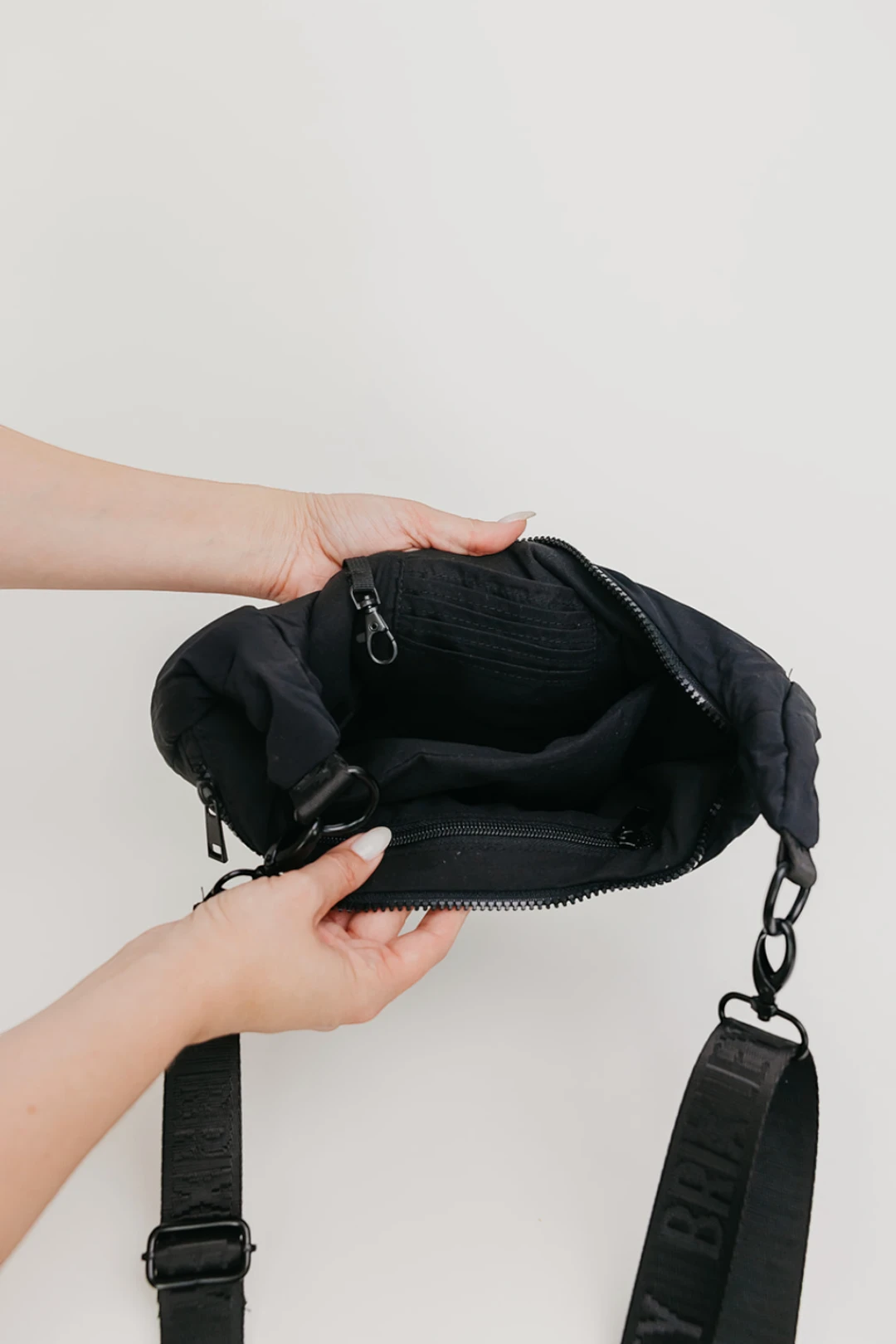 A person's hands holding open a black nylon crossbody bag to show the interior compartments and zipper details, with a black strap hanging down against a plain light gray background.
