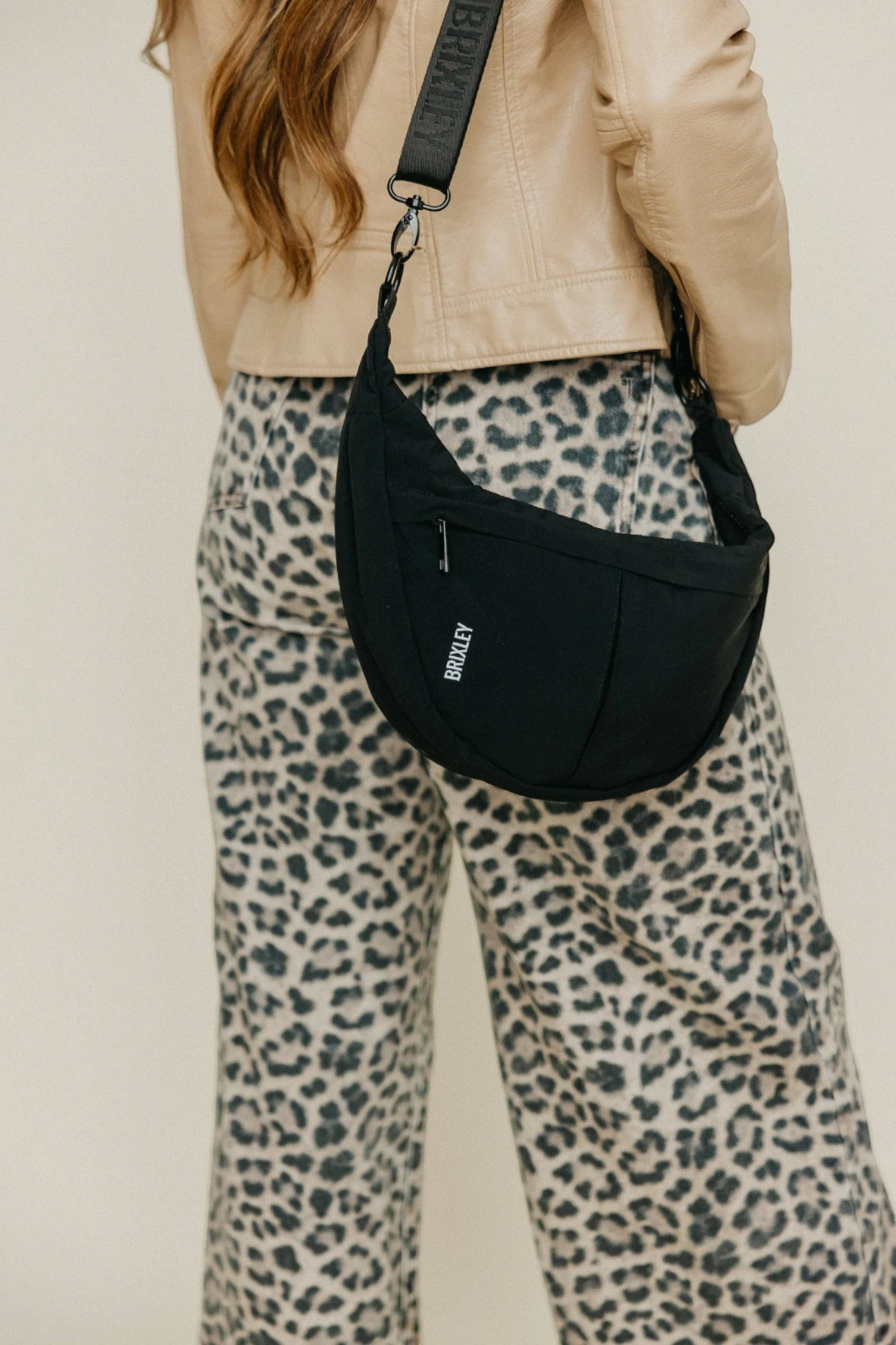 Person wearing a black crescent bag with a beige jacket and leopard print pants on a plain background