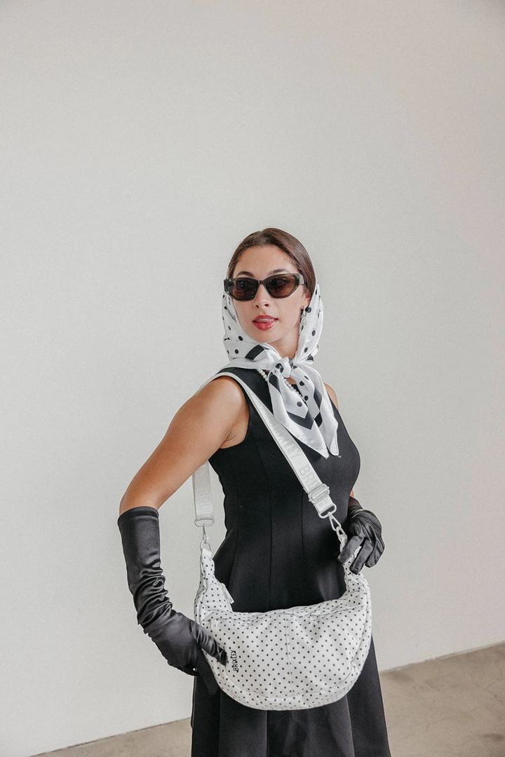 A woman wearing a black dress, black gloves, and a black-and-white polka dot scarf models a matching black-and-white polka dot shoulder bag against a plain light gray wall.