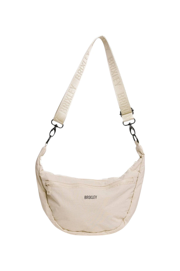 A cream-colored crescent-shaped crossbody bag by BRIXLEY, featuring a matching branded webbing strap and black hardware, shown against a plain white background.