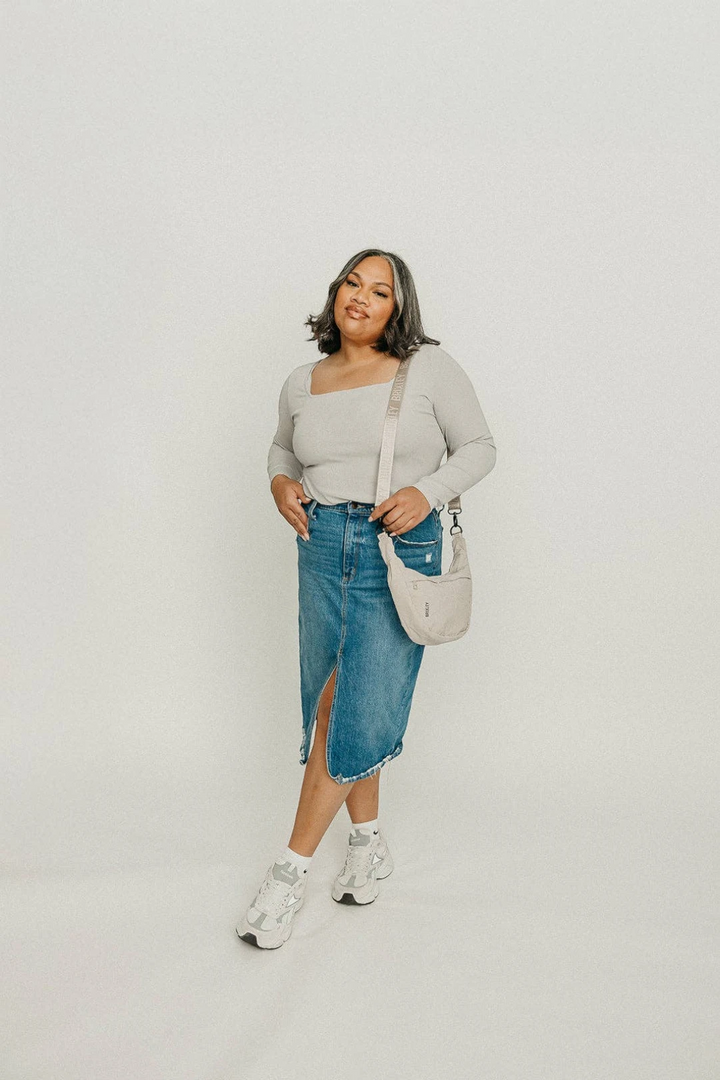 Woman wearing a light gray sweater, blue denim skirt, and white sneakers on a plain background