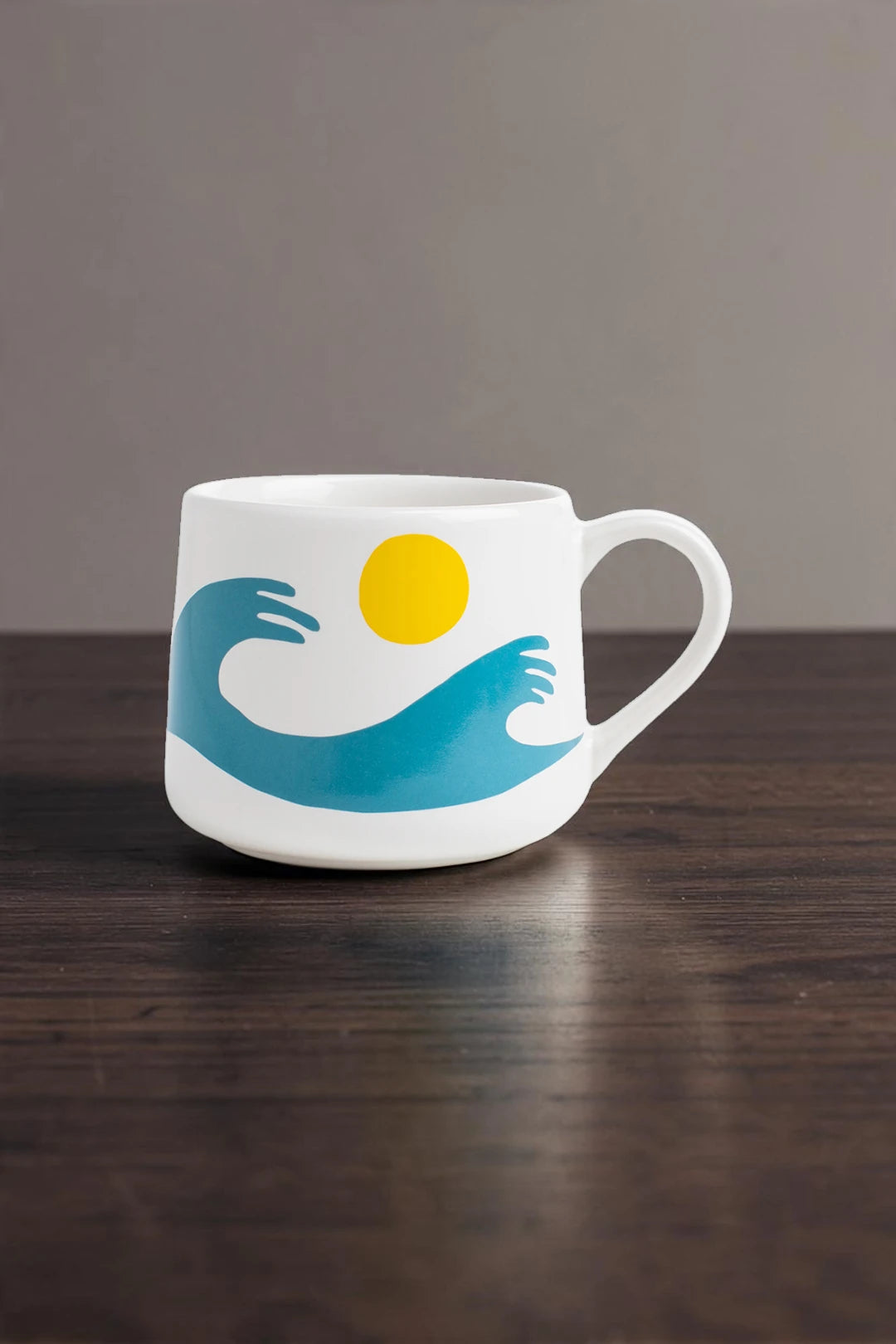 A white ceramic coffee mug with a handle, featuring a graphic design of a blue wave, a yellow sun, and a white cloud, sitting on a dark wooden table against a neutral gray background.
