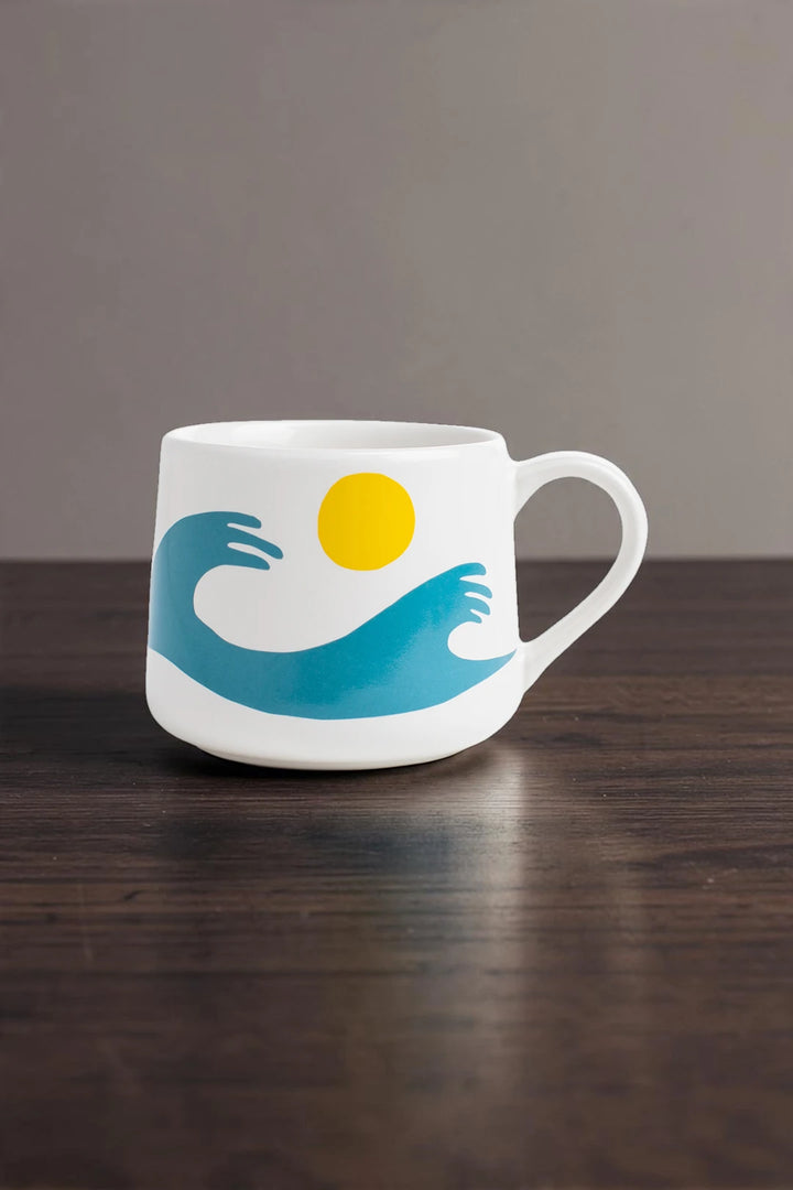 A white ceramic coffee mug with a handle, featuring a graphic design of a blue wave, a yellow sun, and a white cloud, sitting on a dark wooden table against a neutral gray background.