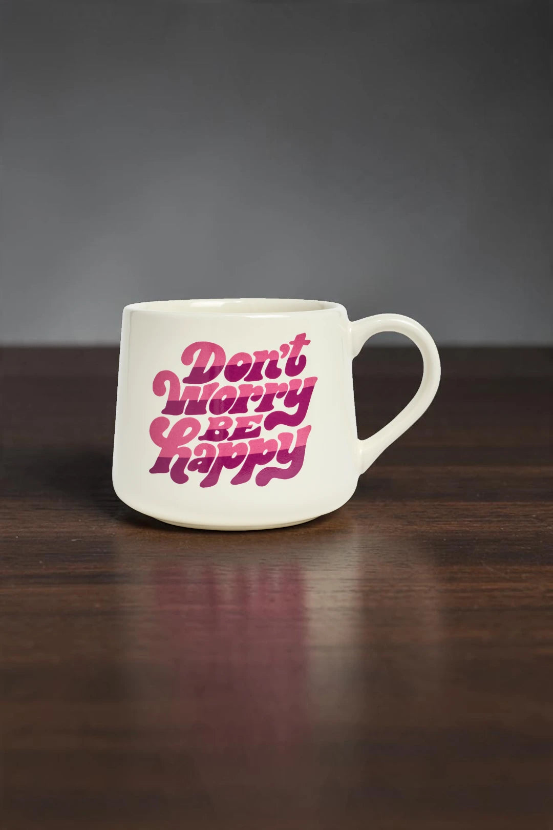A white ceramic coffee mug with the phrase 'Don't Worry Be Happy' printed in a bold pink retro font, resting on a dark wooden surface against a dark gray background.