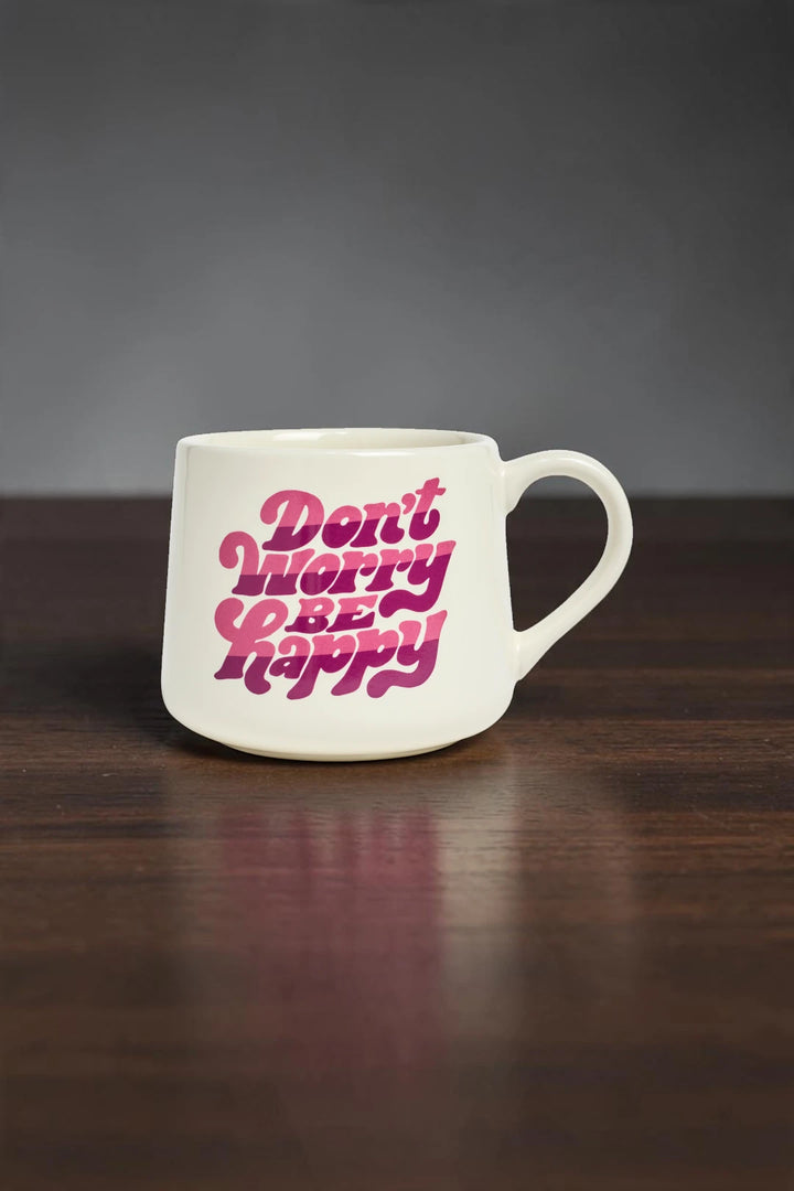 A white ceramic coffee mug with the phrase 'Don't Worry Be Happy' printed in a bold pink retro font, resting on a dark wooden surface against a dark gray background.