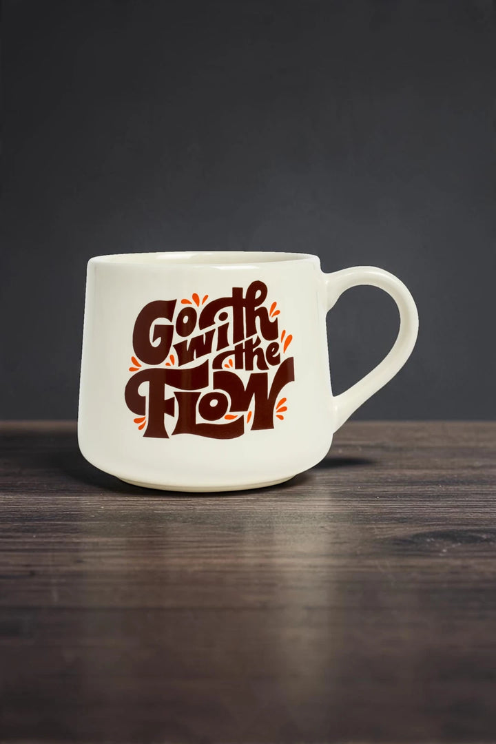 A white ceramic coffee mug with a bold, stylized brown and orange graphic reading 'Go with the Flow' sitting on a dark wooden surface against a dark gray background.