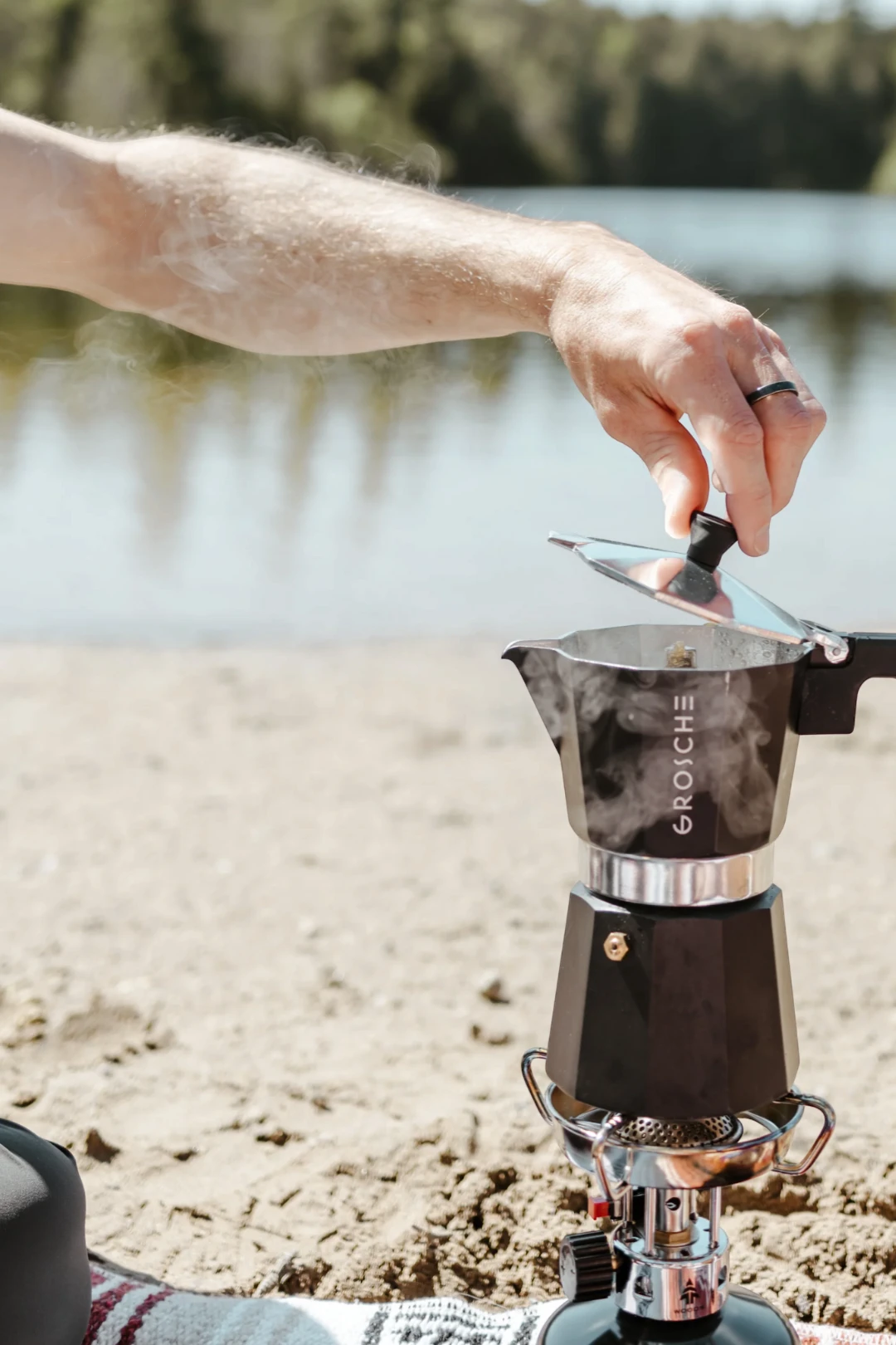 A close-up lifestyle shot of a person's hand lifting the lid of a black and silver Moka pot (espresso maker) set on a portable camping stove outdoors on a sandy shore with water in the background. The brand name 'GROSCHE' is clearly visible on the pot.