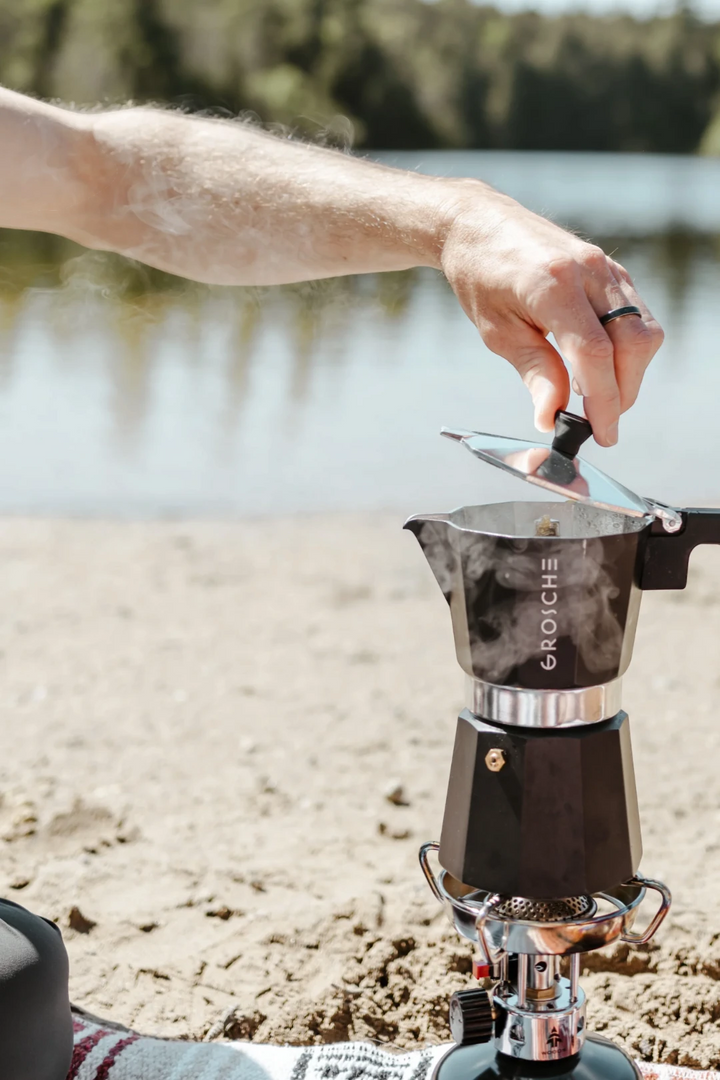 A close-up lifestyle shot of a person's hand lifting the lid of a black and silver Moka pot (espresso maker) set on a portable camping stove outdoors on a sandy shore with water in the background. The brand name 'GROSCHE' is clearly visible on the pot.